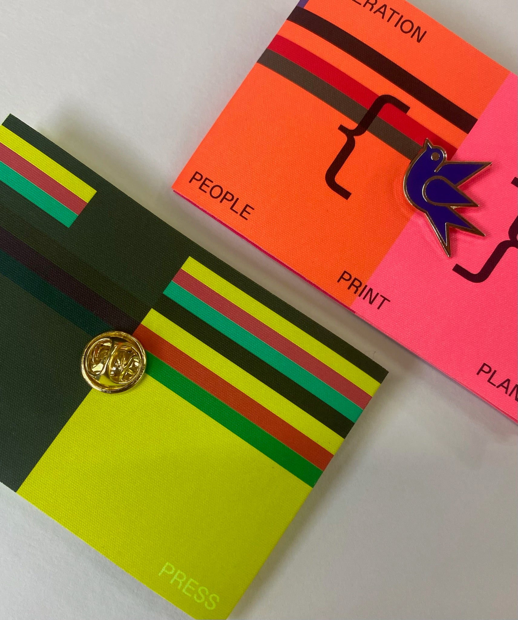 GP Martlet Pin placed on colourful printed cards with bold striped sections and text, arranged on a light background. One card is dark green with yellow, red, and green stripes and a round gold button closure.