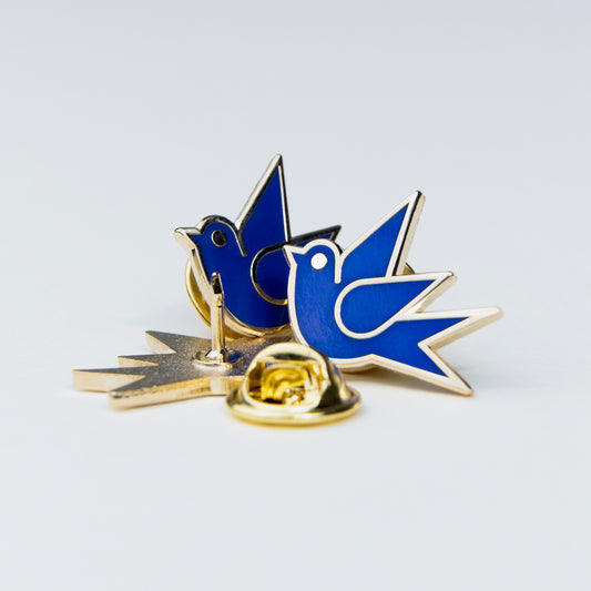 Martlet enamel pin in blue and gold, with one pin showing the back clasp and the other showing the front side, photographed on a clean light gray background.